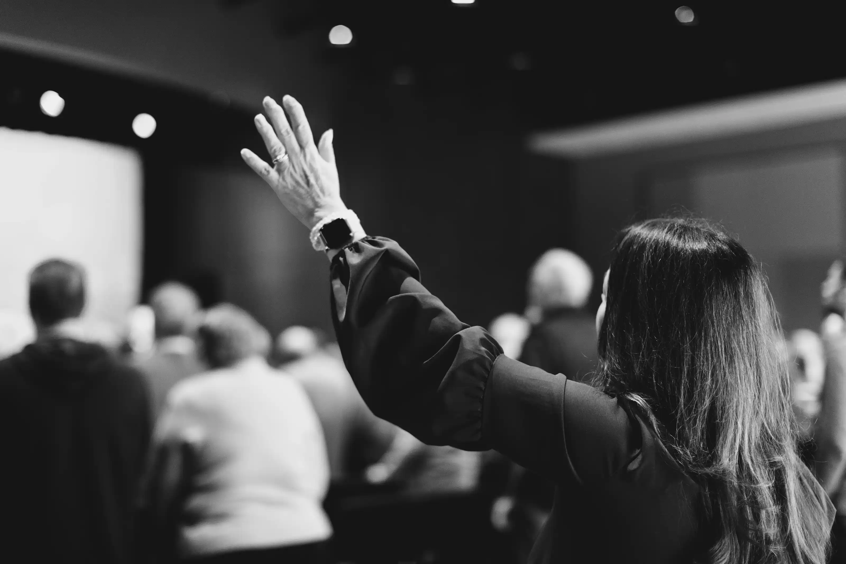 Woman with long hair raising her hand while singing in a gathering of people in a dimly lit room.