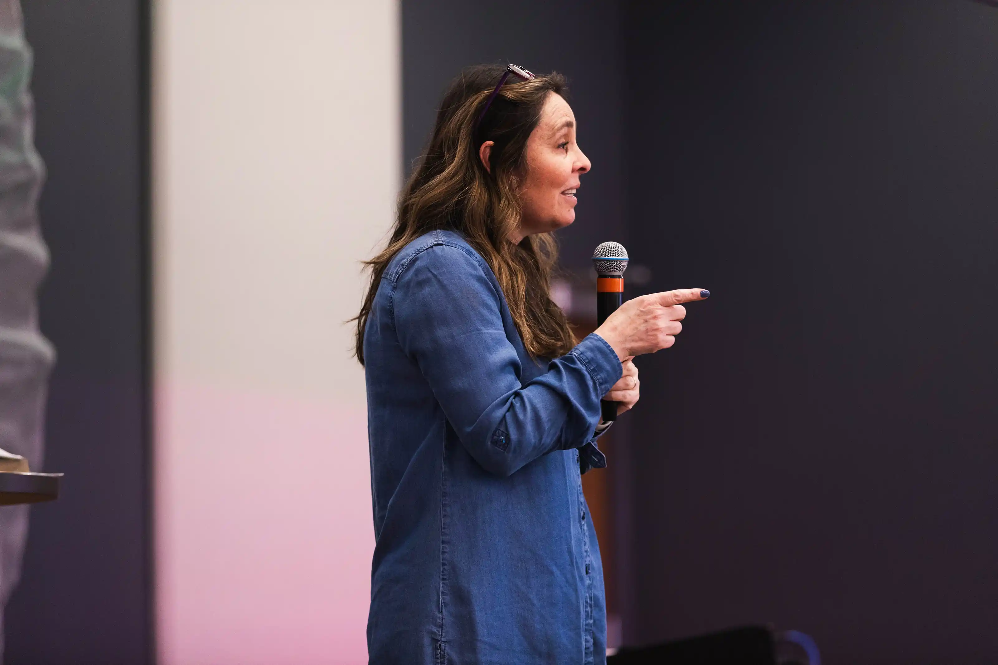 Woman in a blue shirt speaking into a microphone and pointing to her right.