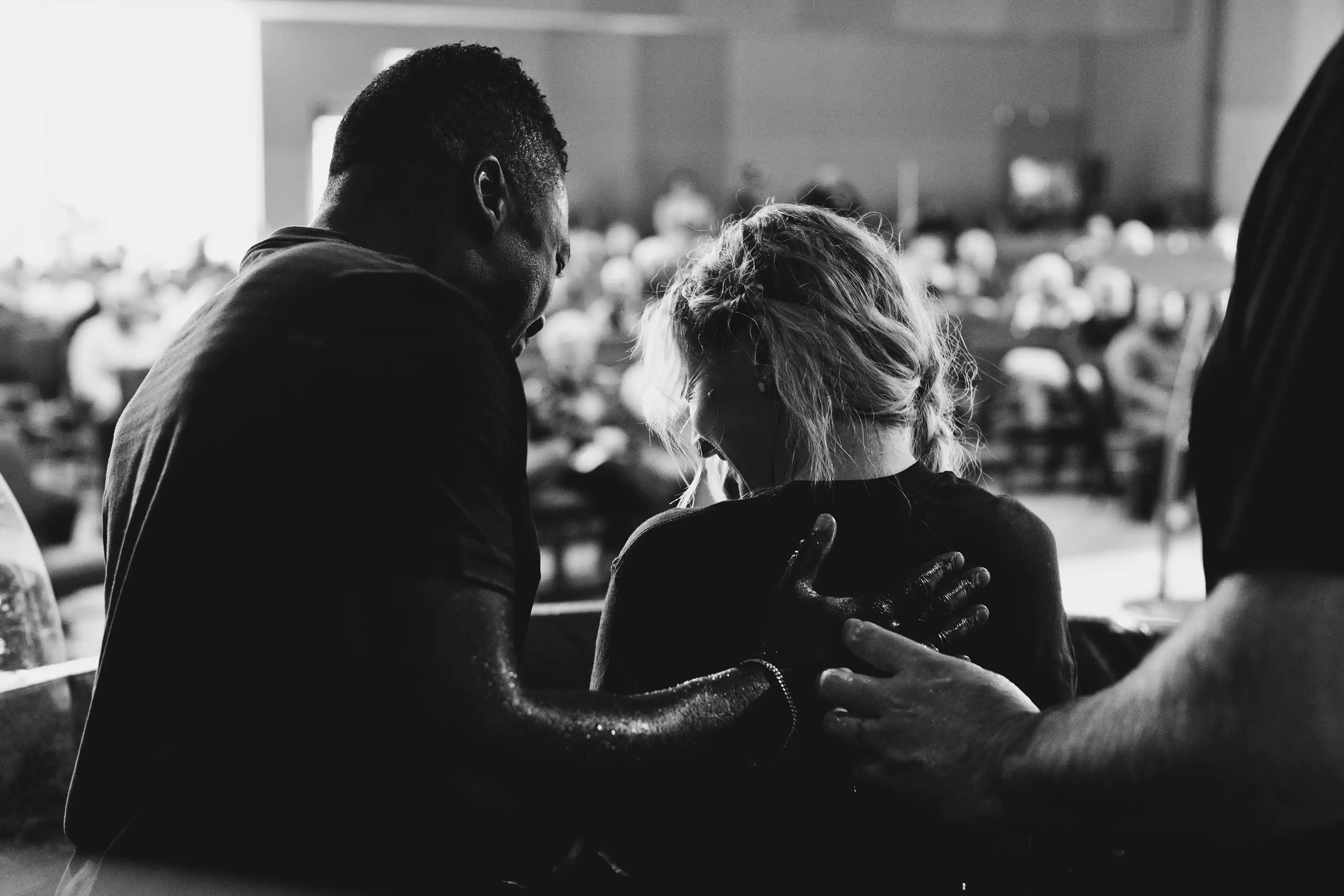 Man with gently placing his hand on a woman's back during a baptism ceremony with an audience in the background.