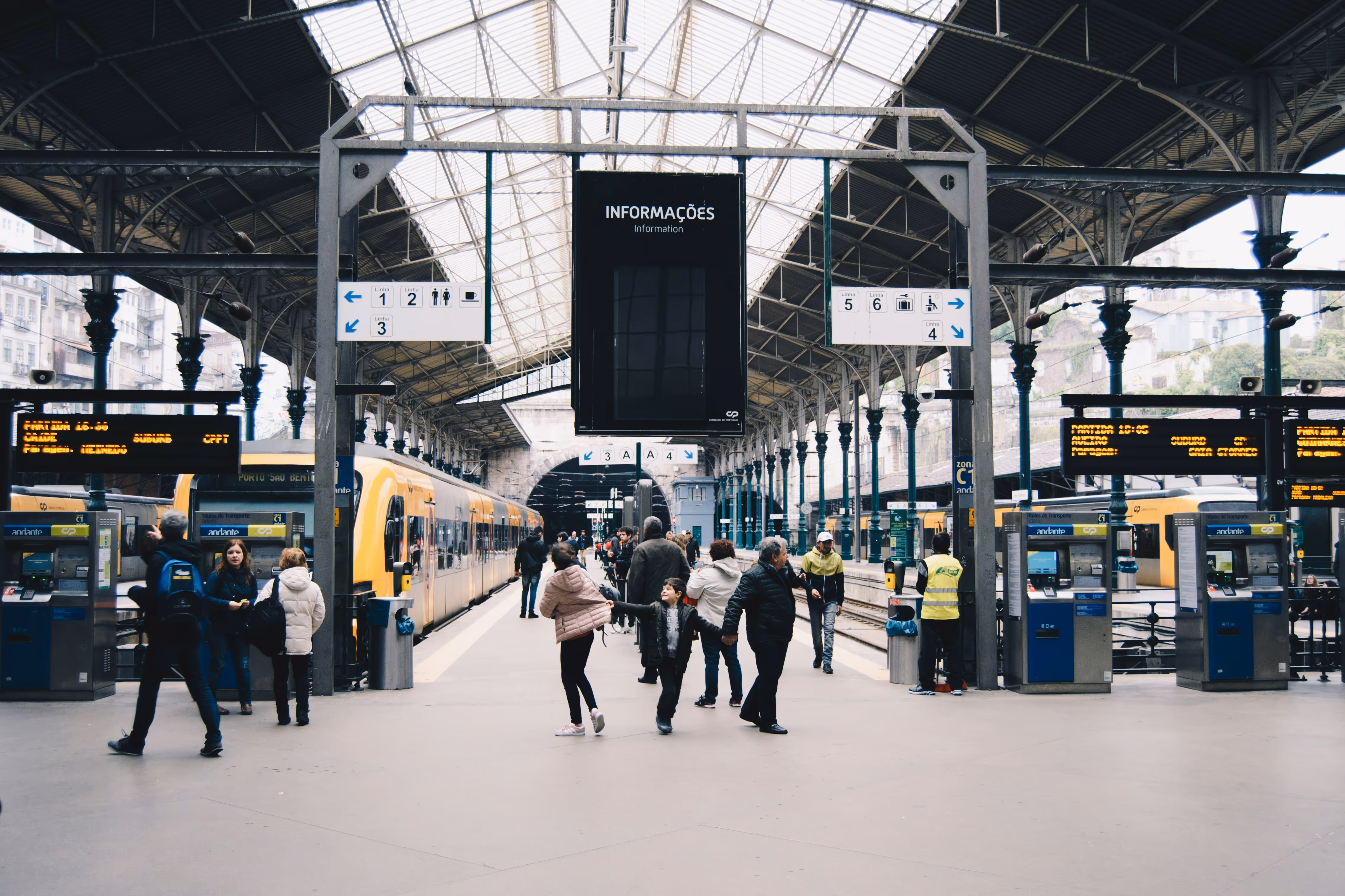 Gare animée avec voyageurs sur le quai, trains jaunes et grand toit vitré métallique.