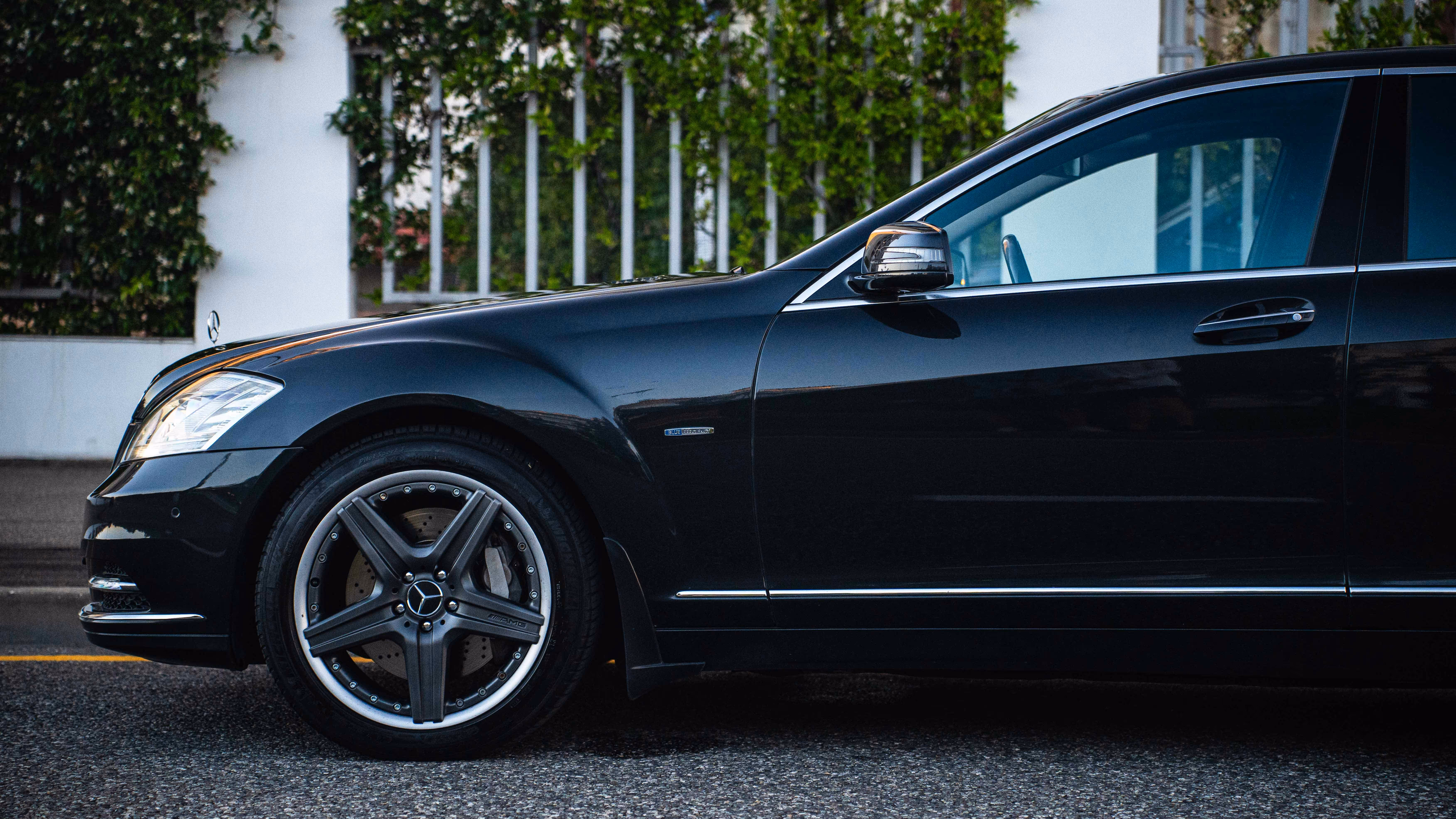 Vue latérale d'une voiture noire Mercedes-Benz garée sur une route goudronnée devant une haie verte.
