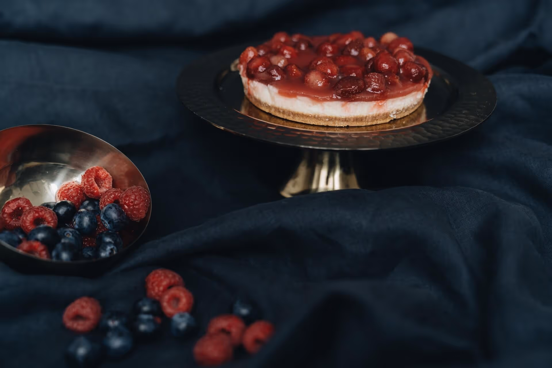 Cheesecake topped with red berries on a gold pedestal with scattered raspberries and blueberries on dark fabric.