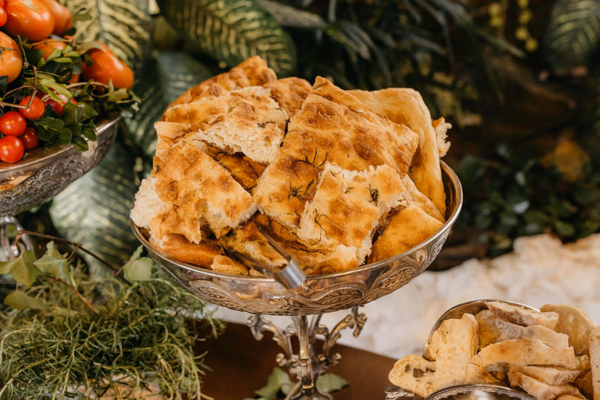 Silver bowl filled with torn focaccia bread pieces, surrounded by fresh herbs and cherry tomatoes.
