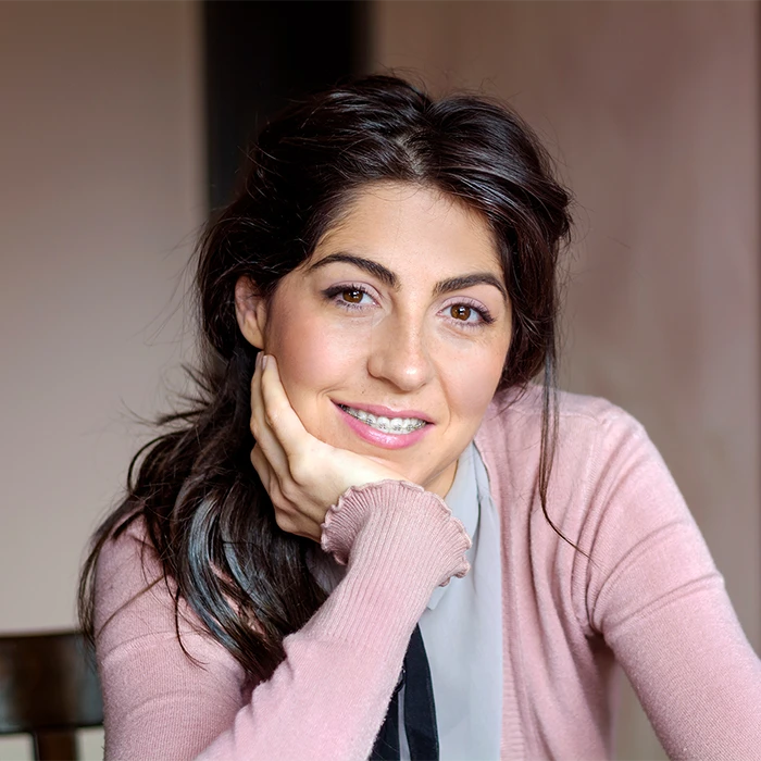 Smiling woman with dark hair wearing braces and a pink sweater resting her chin on her hand.