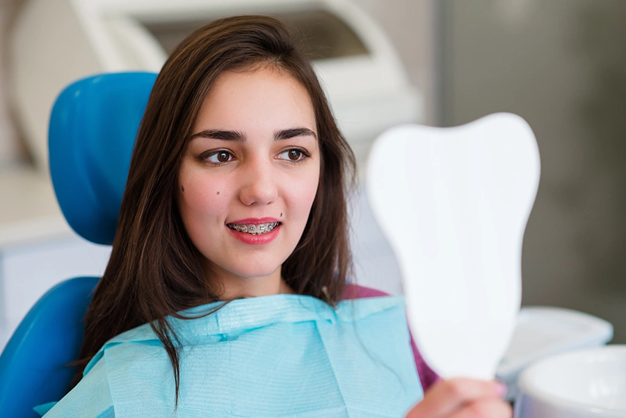Teenage girl with braces sitting in a dental chair and looking at a tooth-shaped mirror.