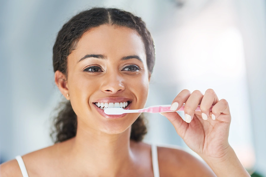 Smiling woman brushing her teeth with a pink toothbrush in a bright bathroom.