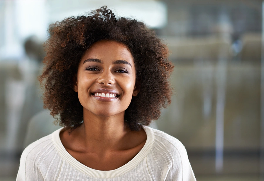 Smiling young woman with curly hair wearing a white sweater against a blurred background.