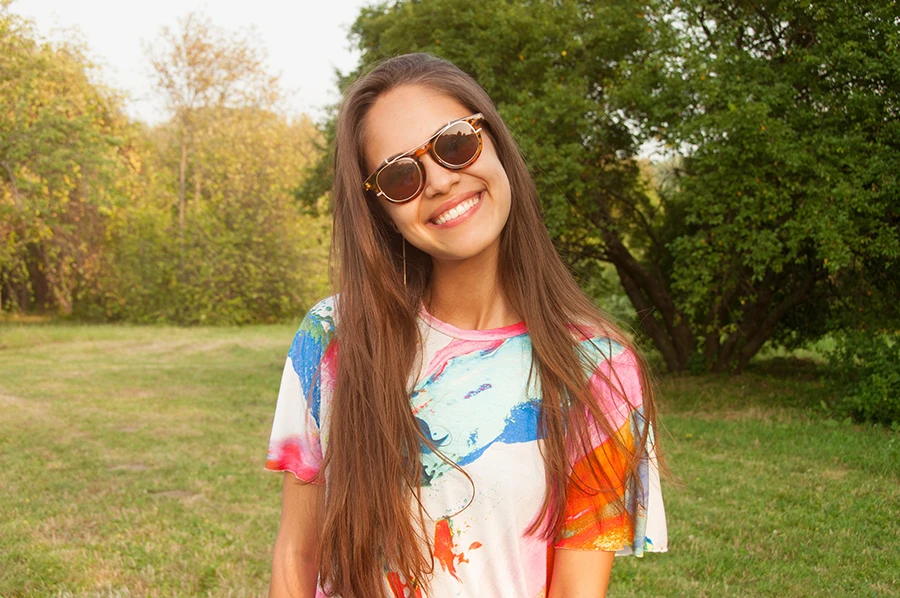 Smiling young woman with long brown hair wearing sunglasses and a colorful tie-dye shirt standing outdoors in a grassy area with trees.
