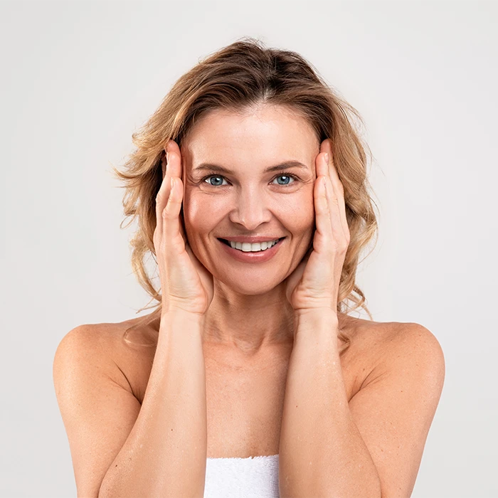 Smiling middle-aged woman with blue eyes and wavy hair gently touching her face with both hands.