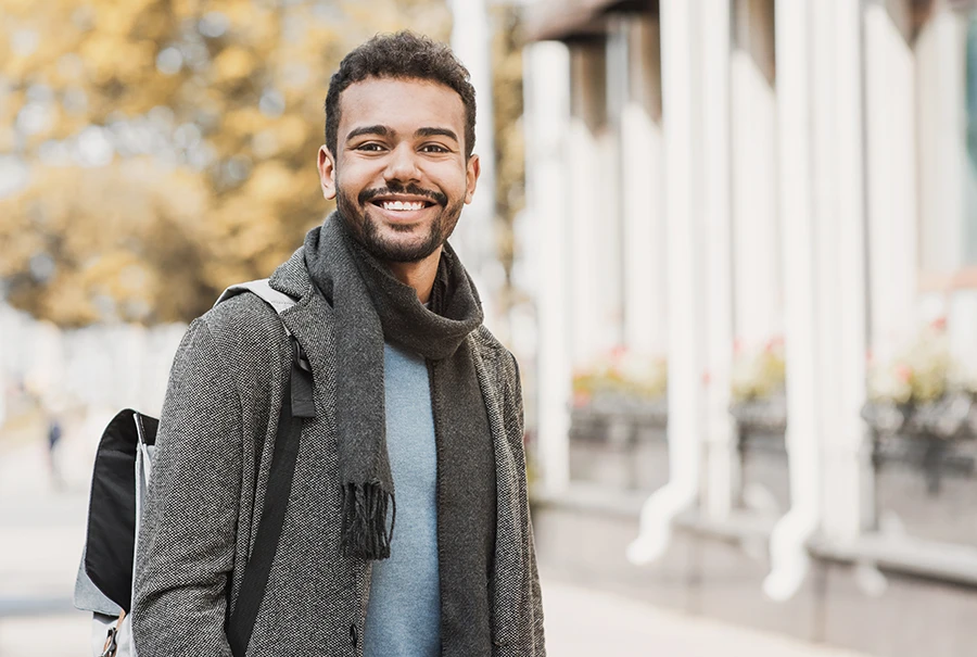 Smiling young man wearing a gray coat and scarf with a backpack walking in a city street.