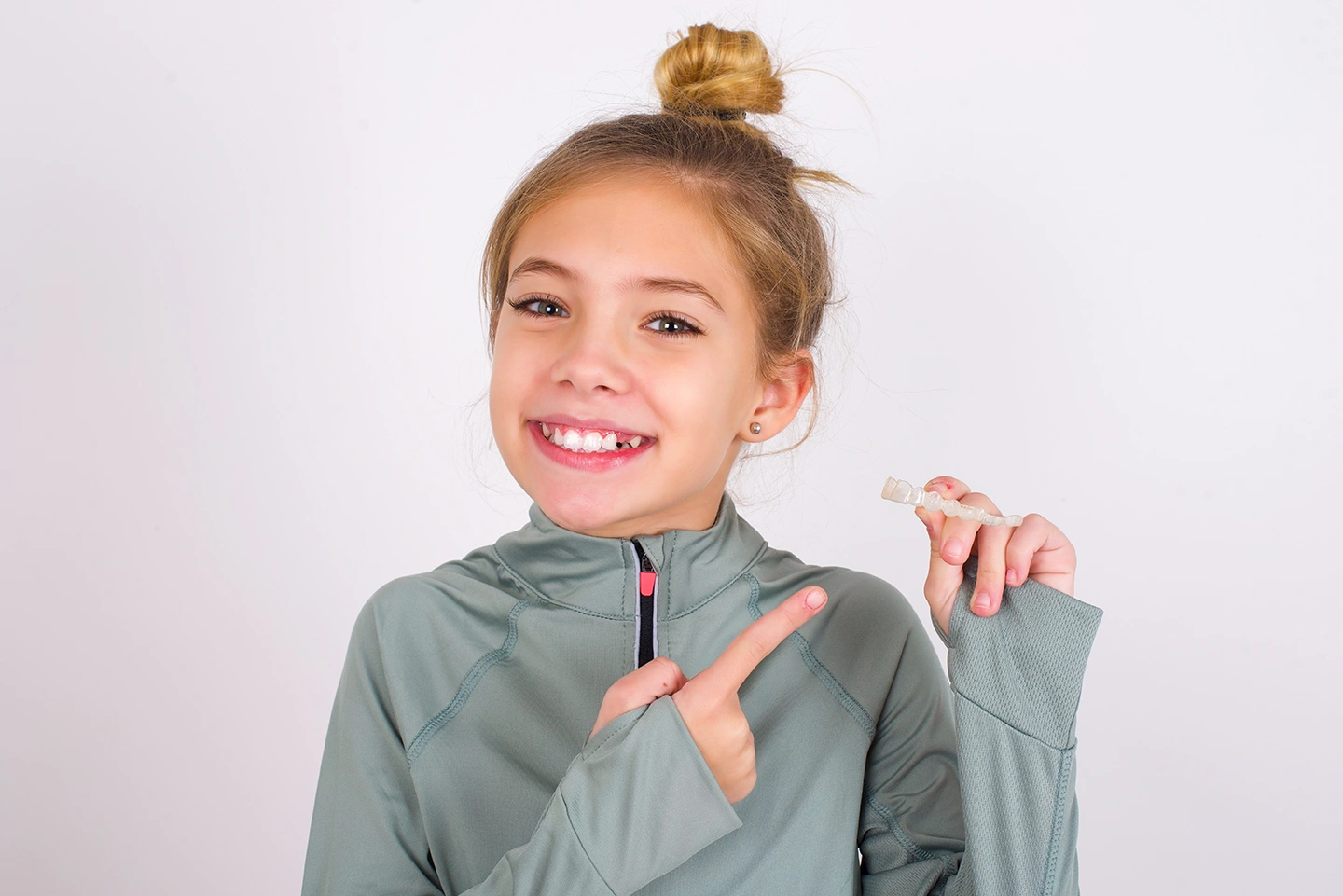 Smiling young girl with a top bun hairstyle pointing at a clear dental aligner she is holding in her hand.