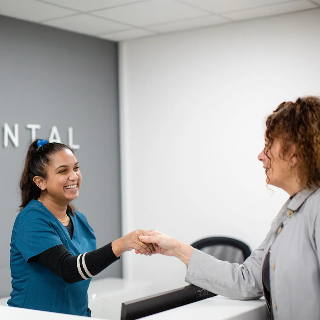 Smiling dental receptionist shaking hands with a woman at the front desk.