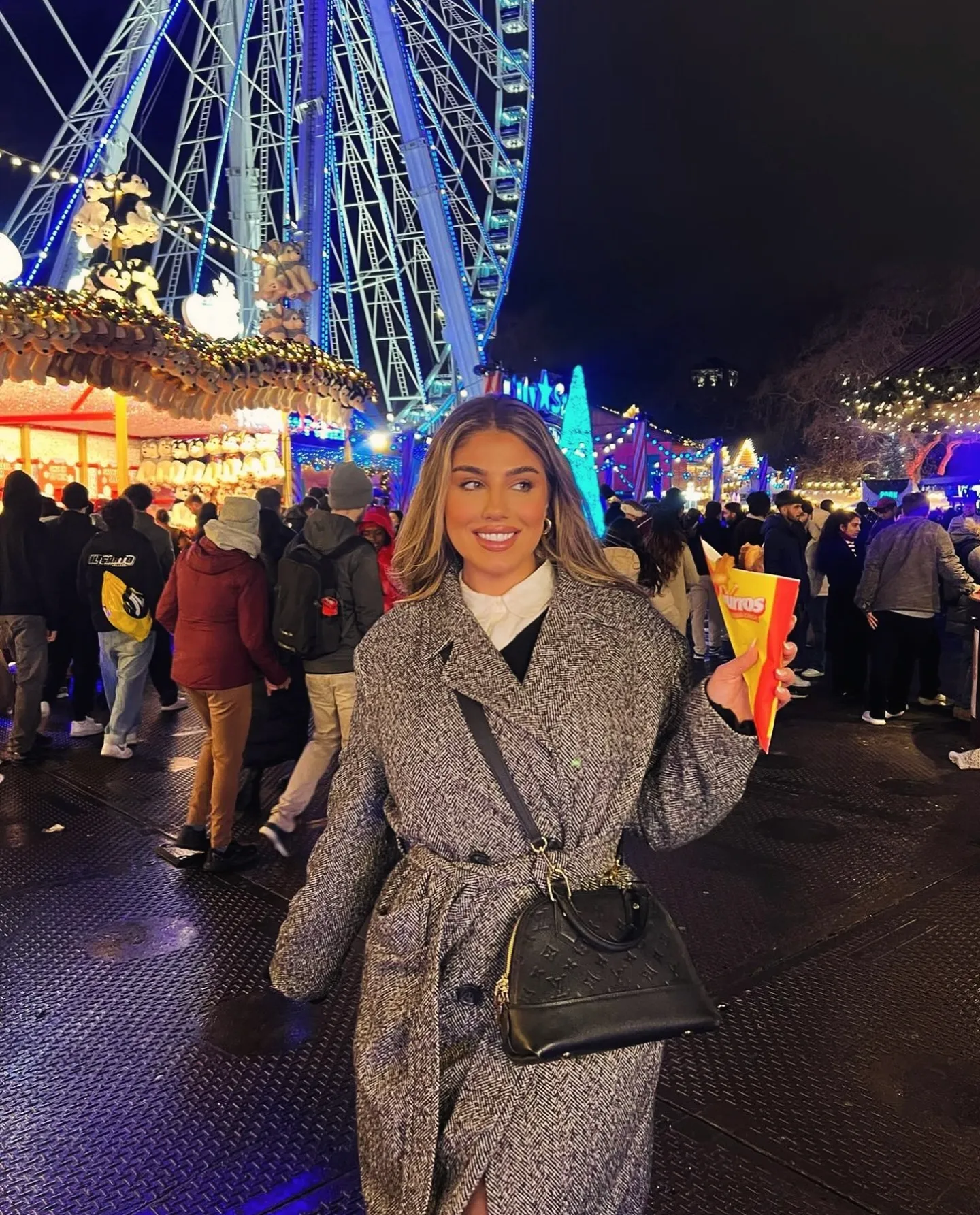 Smiling woman in a gray coat holding churros at a lively nighttime fair with a ferris wheel and game booth lights in the background.