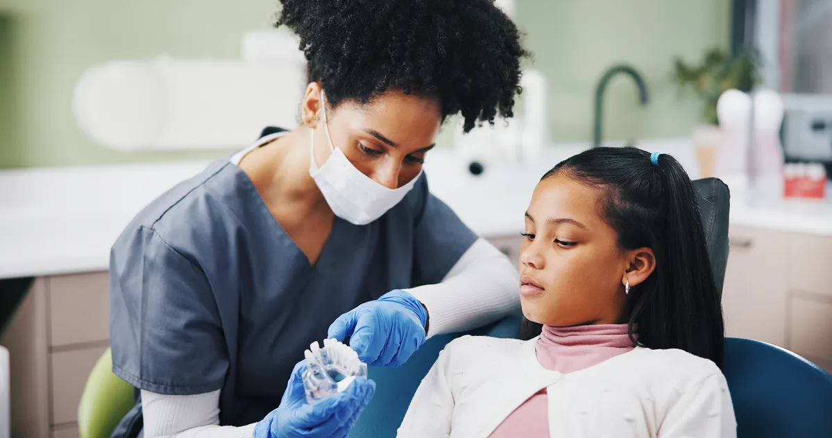 Female dentist wearing a mask and gloves showing a dental shade guide to a young girl in a dental chair.