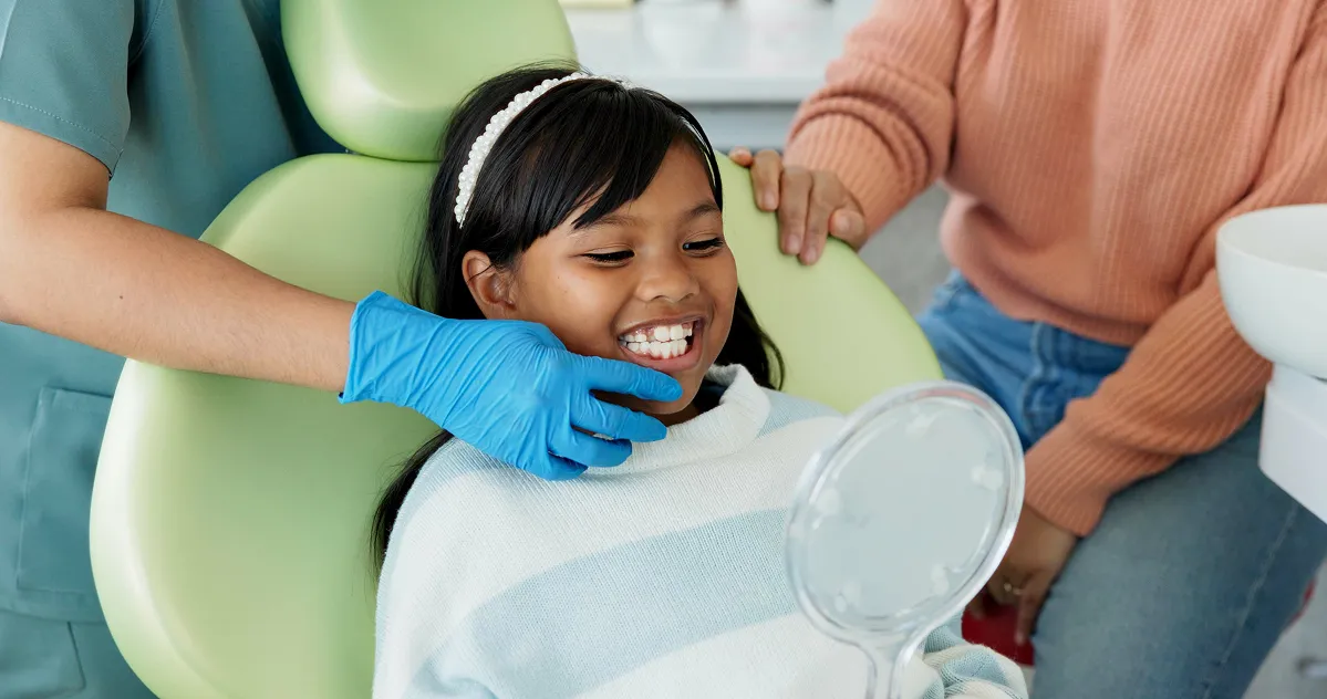 Smiling young girl in dental chair looking at mirror while dentist in blue gloves touches her chin and adult supports her from behind.