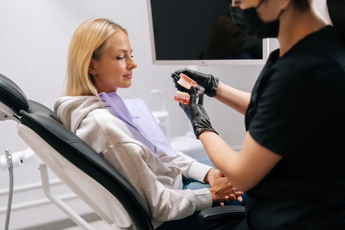 Dentist wearing black gloves shows dental model to a seated patient in a dental clinic.