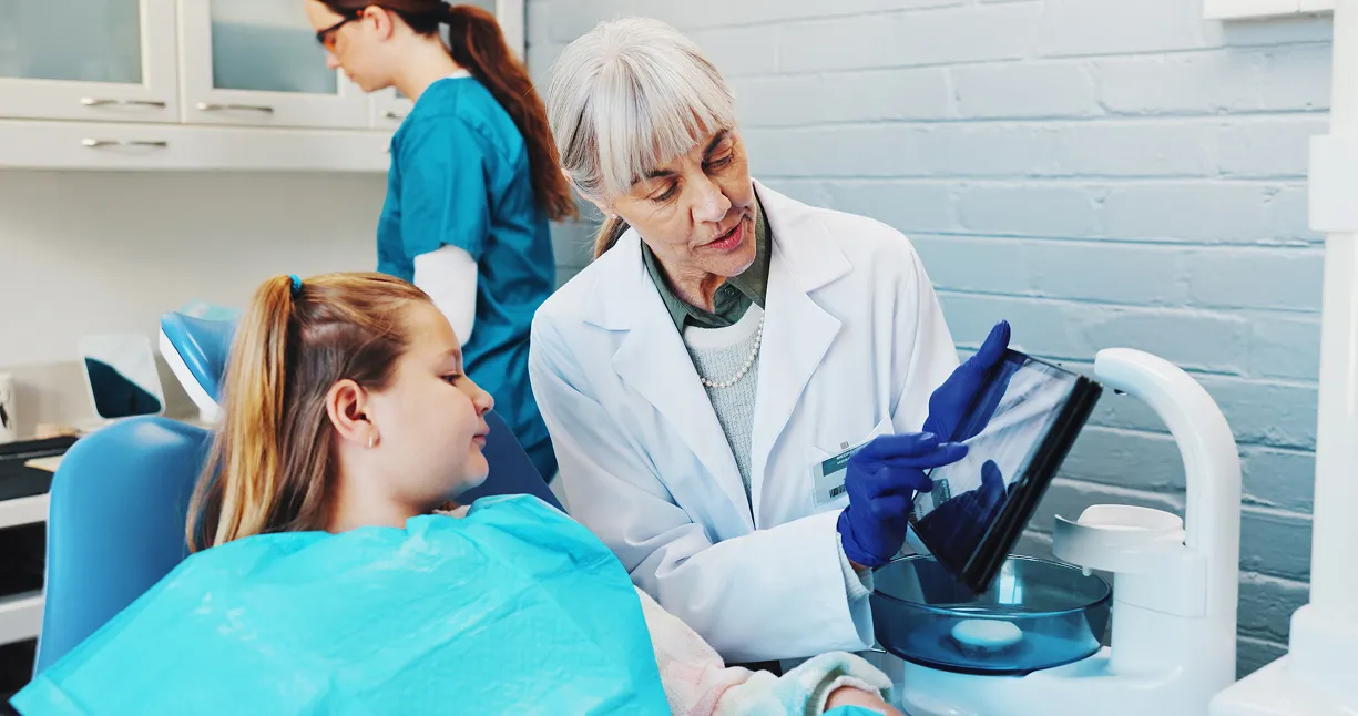 Dentist in white coat showing dental X-ray to a young girl patient in a dental chair while a dental assistant works in the background.
