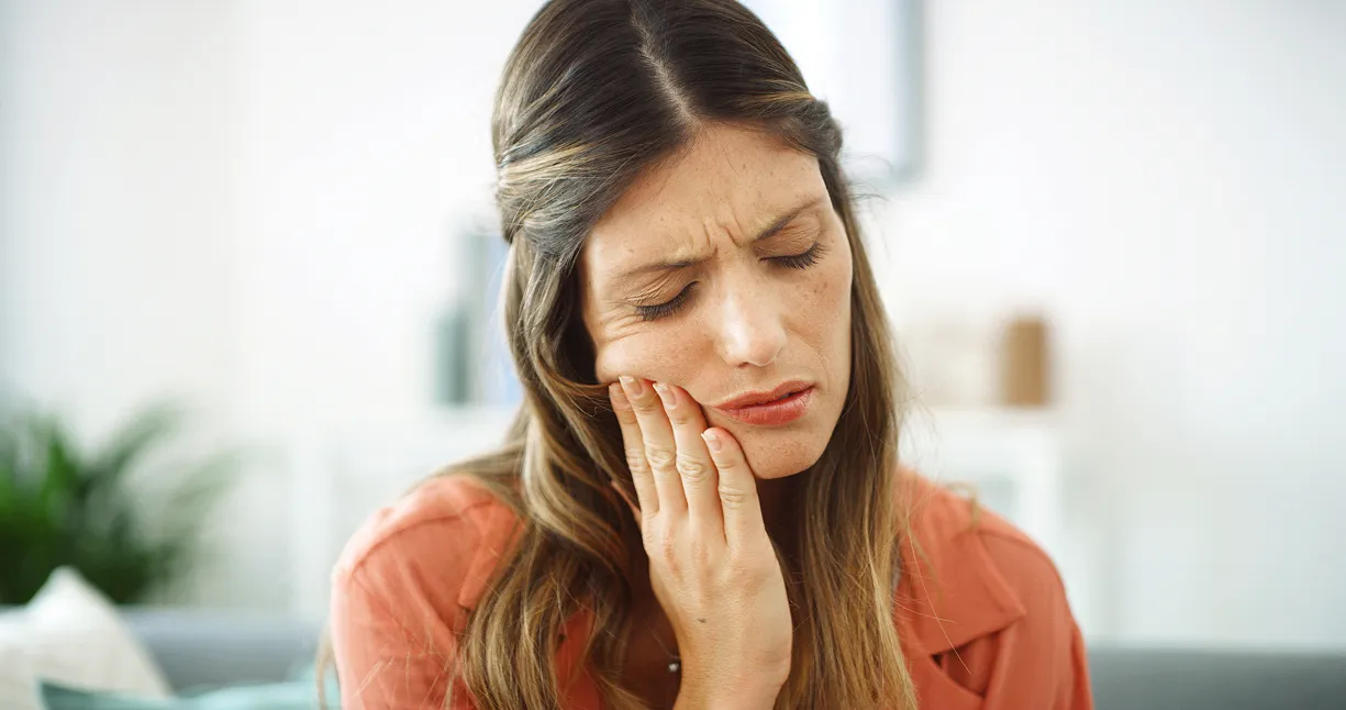 Woman with long brown hair wearing an orange shirt, holding her cheek in pain with eyes closed.