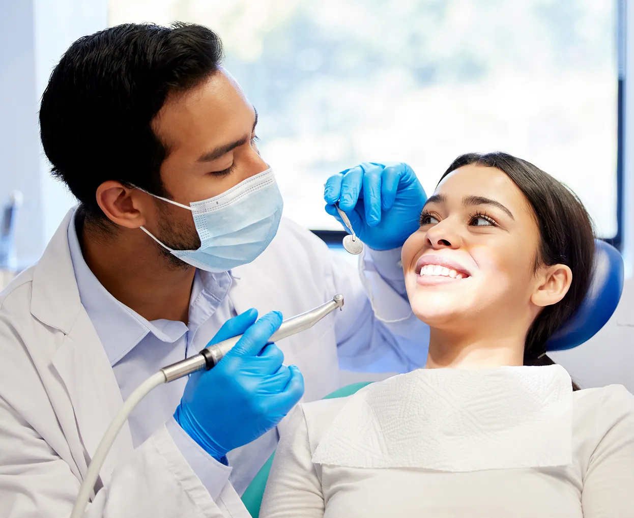 Dentist wearing a mask and blue gloves examining a smiling woman's teeth with dental tools in a clinic.