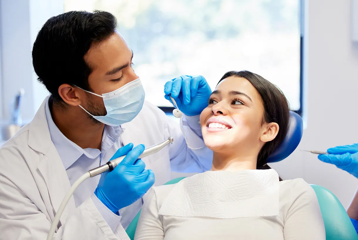 Dentist wearing a mask and blue gloves examining a smiling female patient's teeth in a dental clinic.
