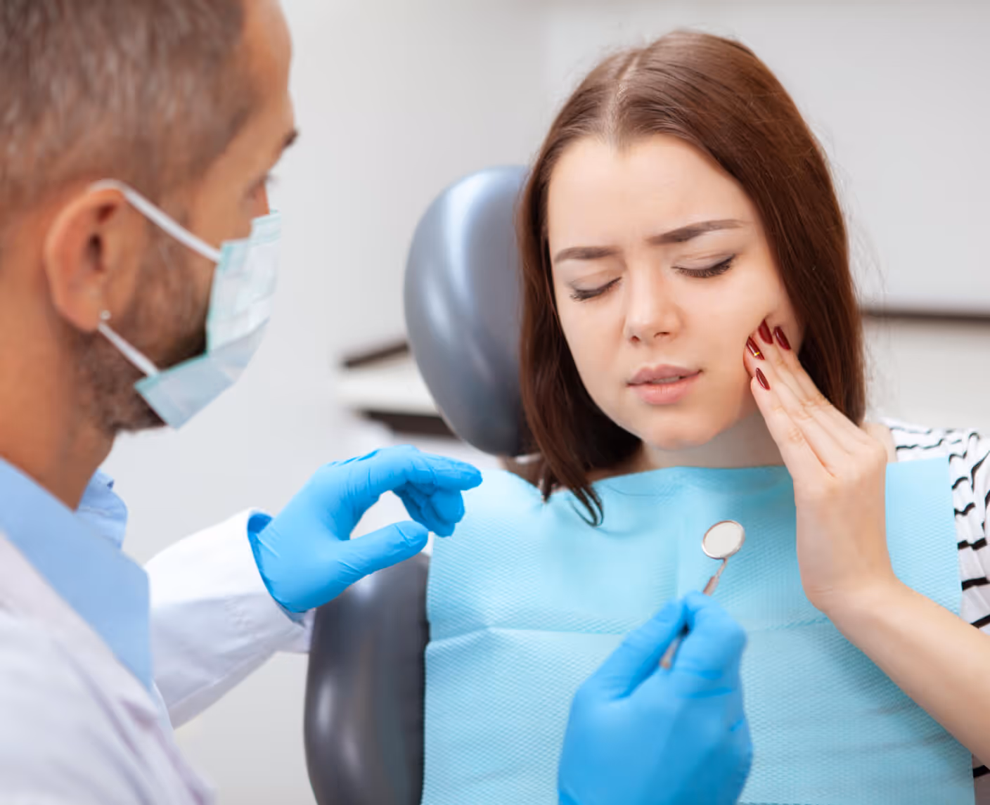 Woman in dental chair holding her cheek in pain while dentist wearing mask and gloves examines her with a dental mirror.