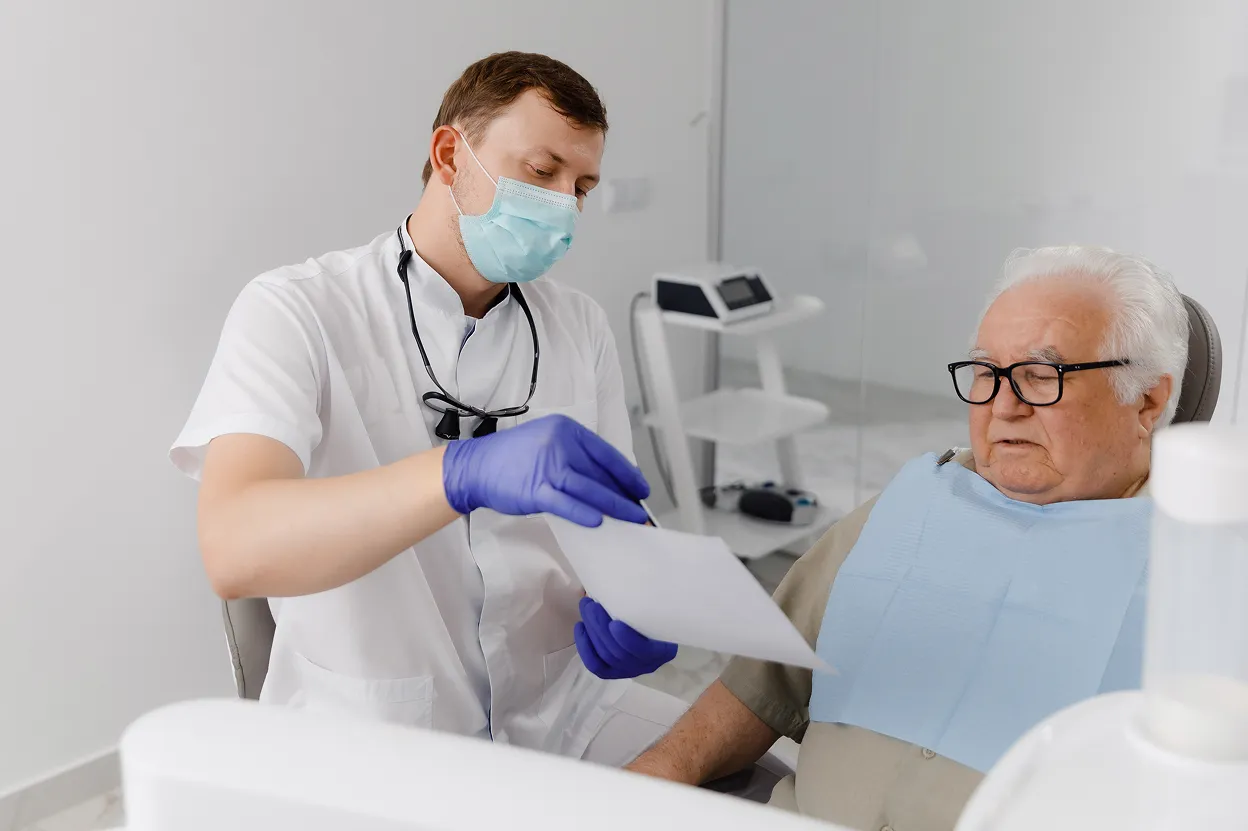 Dentist in a mask and gloves showing a document to an elderly male patient sitting in a dental chair.