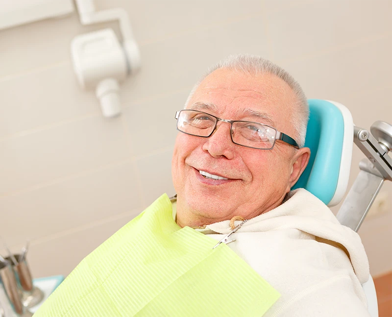 Elderly man wearing glasses smiling while sitting in a dental chair with a dental bib.