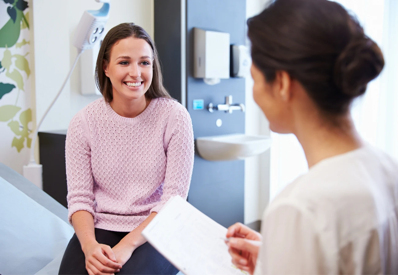 A woman in a pink sweater sitting and smiling during a consultation with a healthcare professional holding a clipboard.