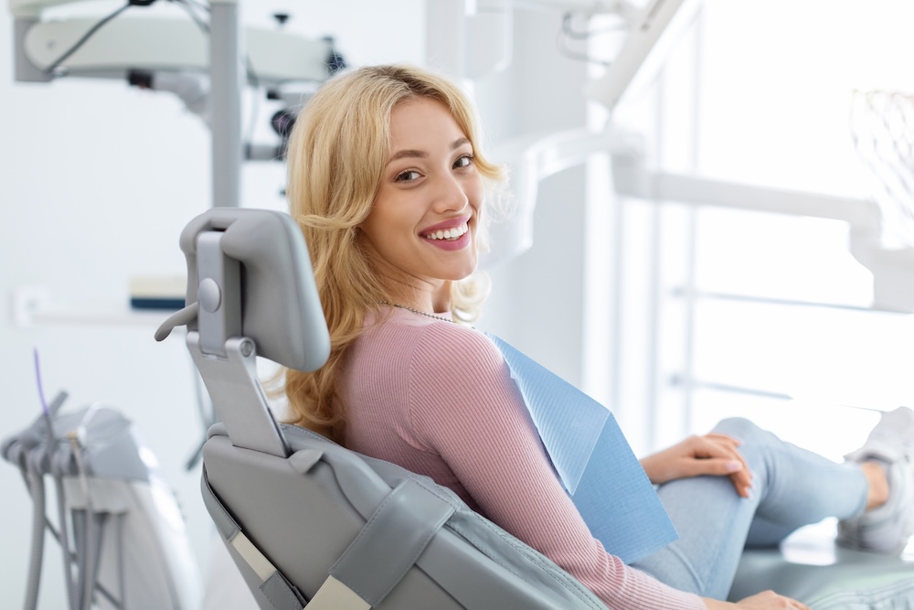 Smiling woman with blonde hair reclining in a dental chair wearing a blue dental bib.