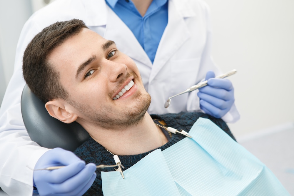 Smiling young man reclining in a dental chair with a dentist holding dental tools behind him.