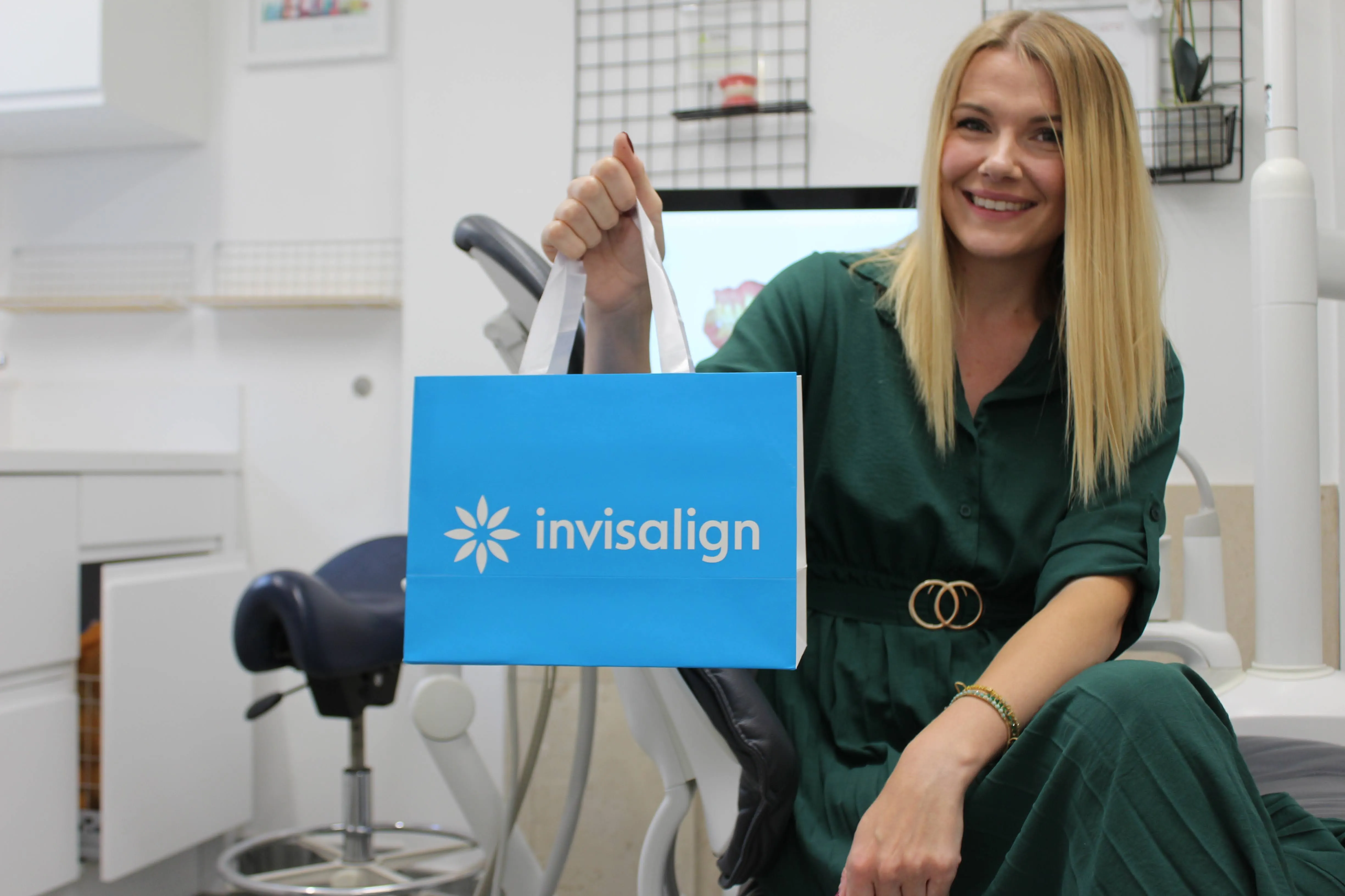 Smiling woman in a green dress sitting in a dental office holding a blue Invisalign bag.