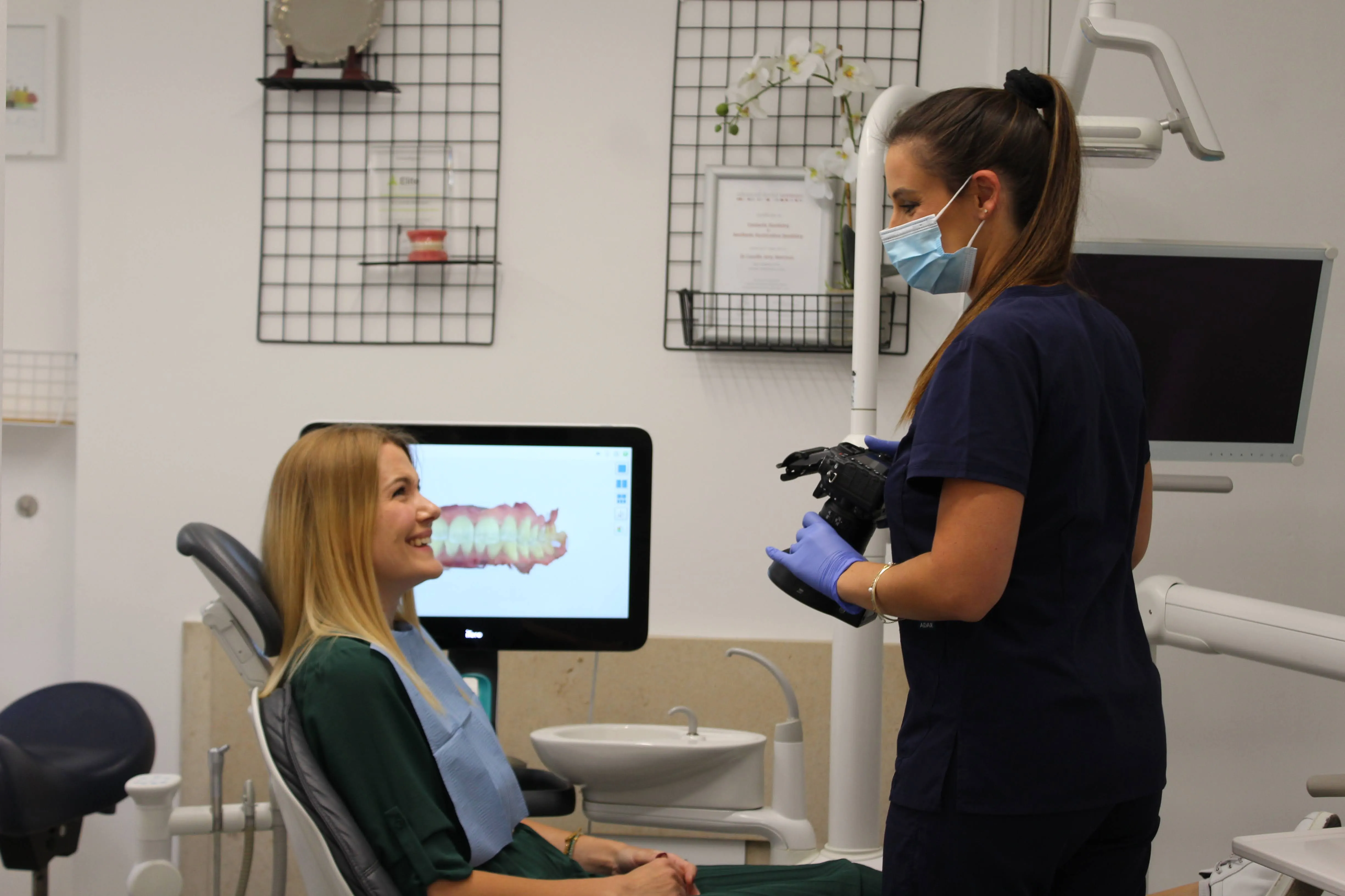 Dental professional wearing a mask holds a camera while talking to a smiling patient seated in a dental chair, with a digital image of teeth displayed on a monitor behind them.