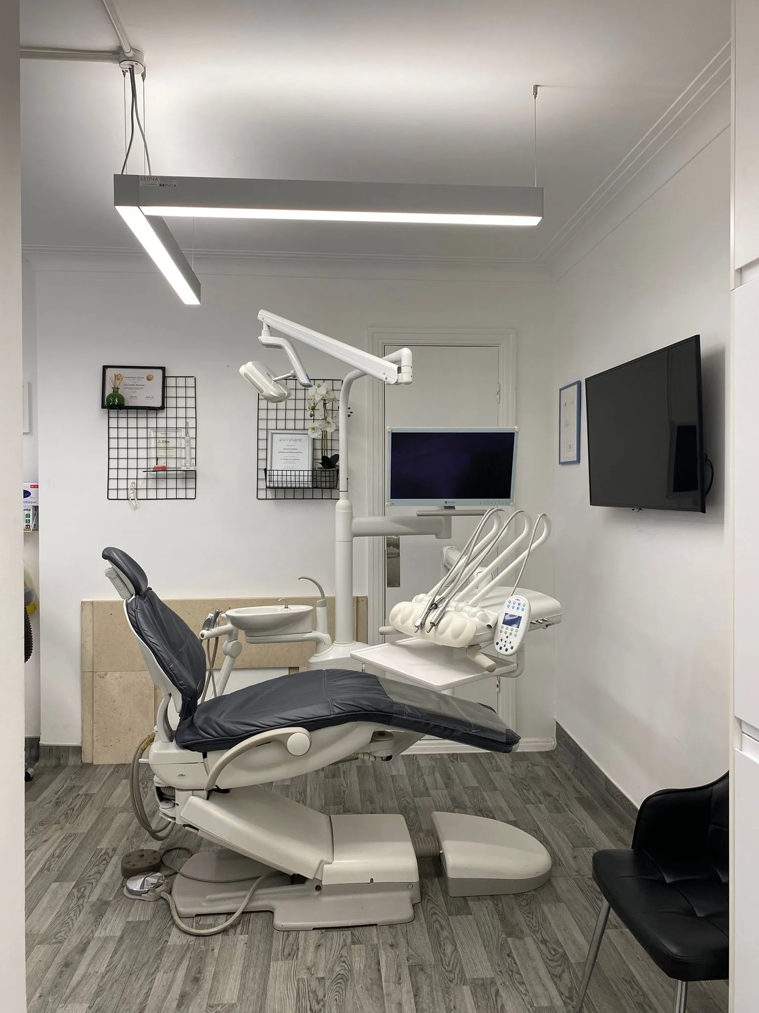 Modern dental examination room with a reclining dental chair, overhead light, instrument tray, two wall-mounted screens, and a black visitor chair.