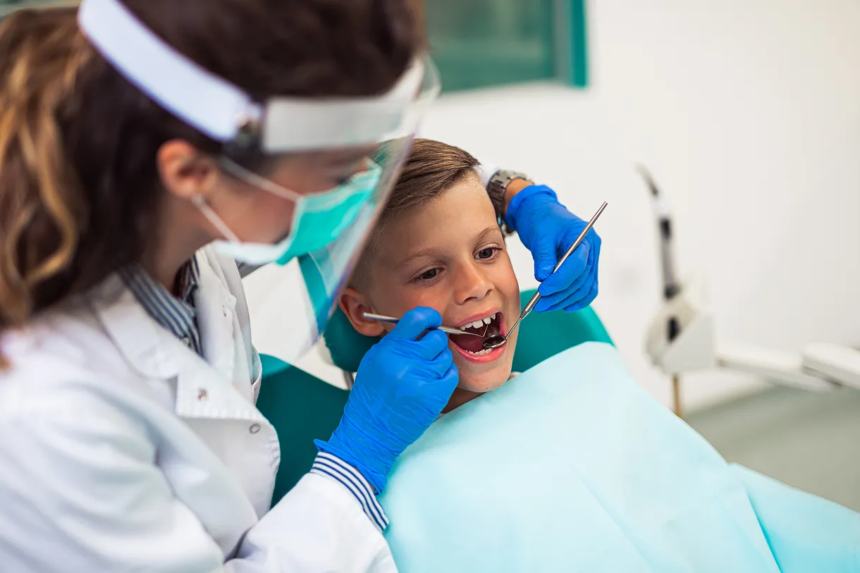 Dentist wearing gloves and a face shield examining the open mouth of a young boy sitting in a dental chair.