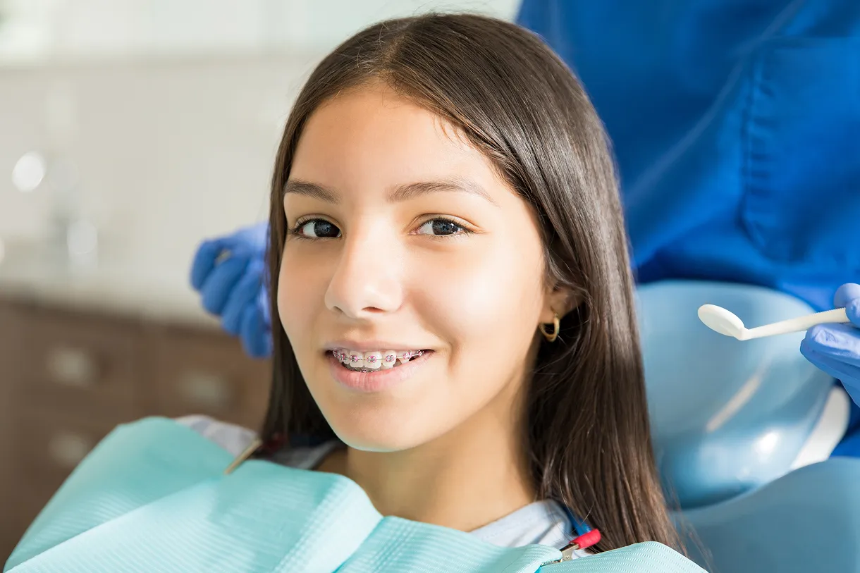Smiling girl with braces sitting in a dental chair wearing a protective bib with a dentist holding a dental tool in the background.