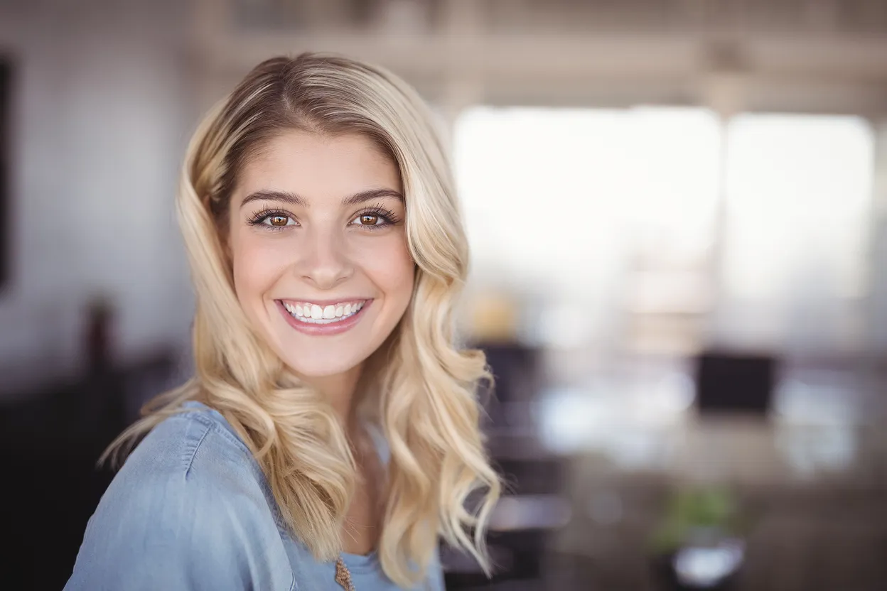 Smiling young woman with long blonde hair and a light blue shirt in a bright indoor setting.