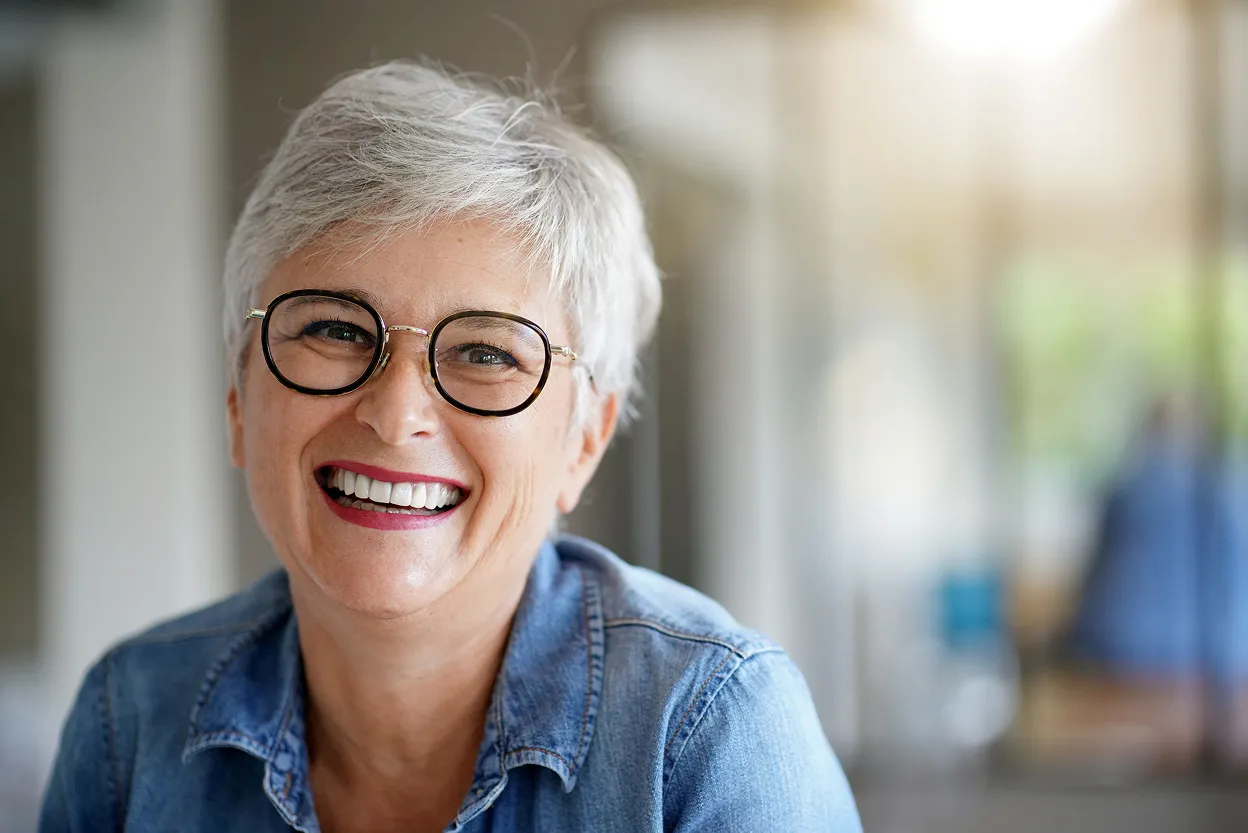 Smiling older woman with short gray hair and round glasses wearing a denim shirt.