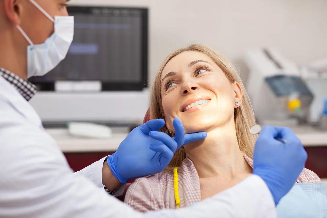 Dentist wearing blue gloves examines a smiling female patient's teeth in a dental office.