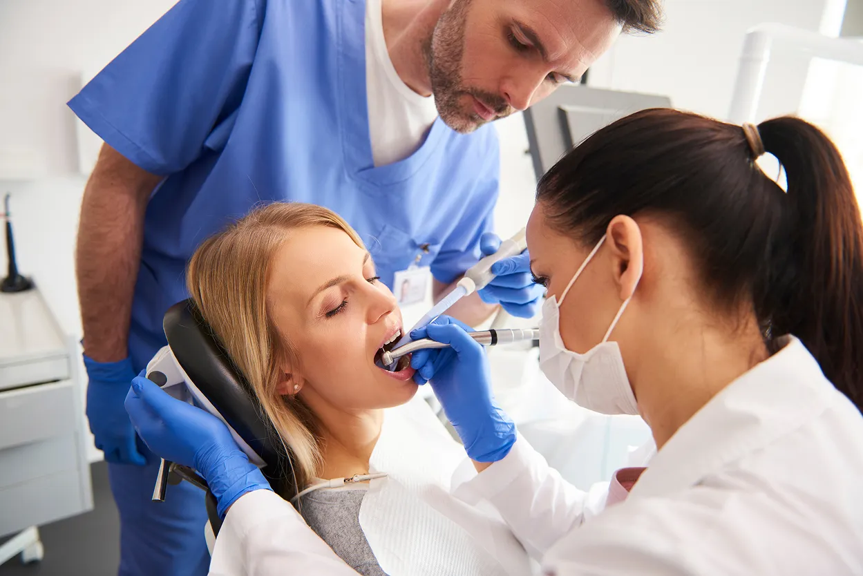 Dentist wearing gloves and mask examining a female patient's mouth with a dental tool as an assistant observes.