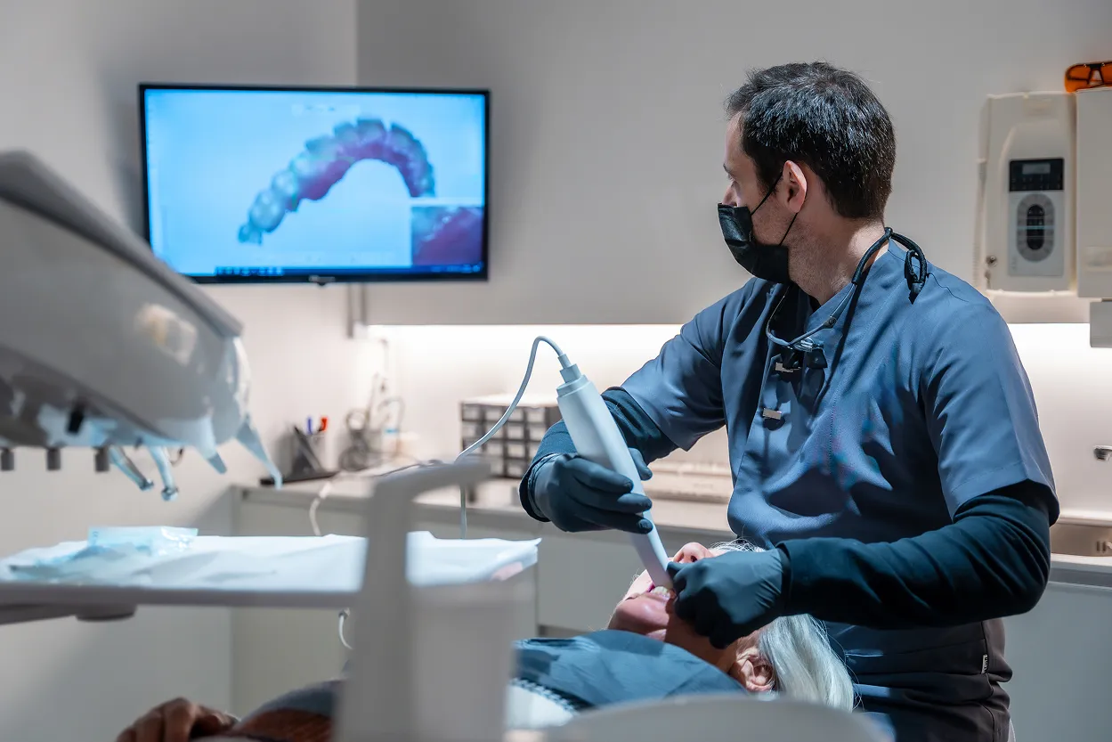 Dentist wearing black mask and gloves using a dental scanner on a patient in a modern clinic with a dental image displayed on a monitor.