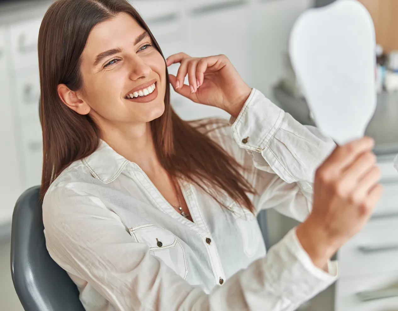 Smiling young woman in a white shirt looking at her reflection in a handheld mirror.