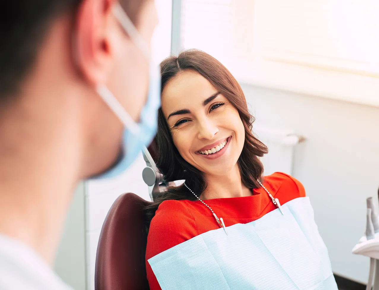 Smiling woman in a red top sitting in a dental chair wearing a dental bib, looking at a dentist with a mask.