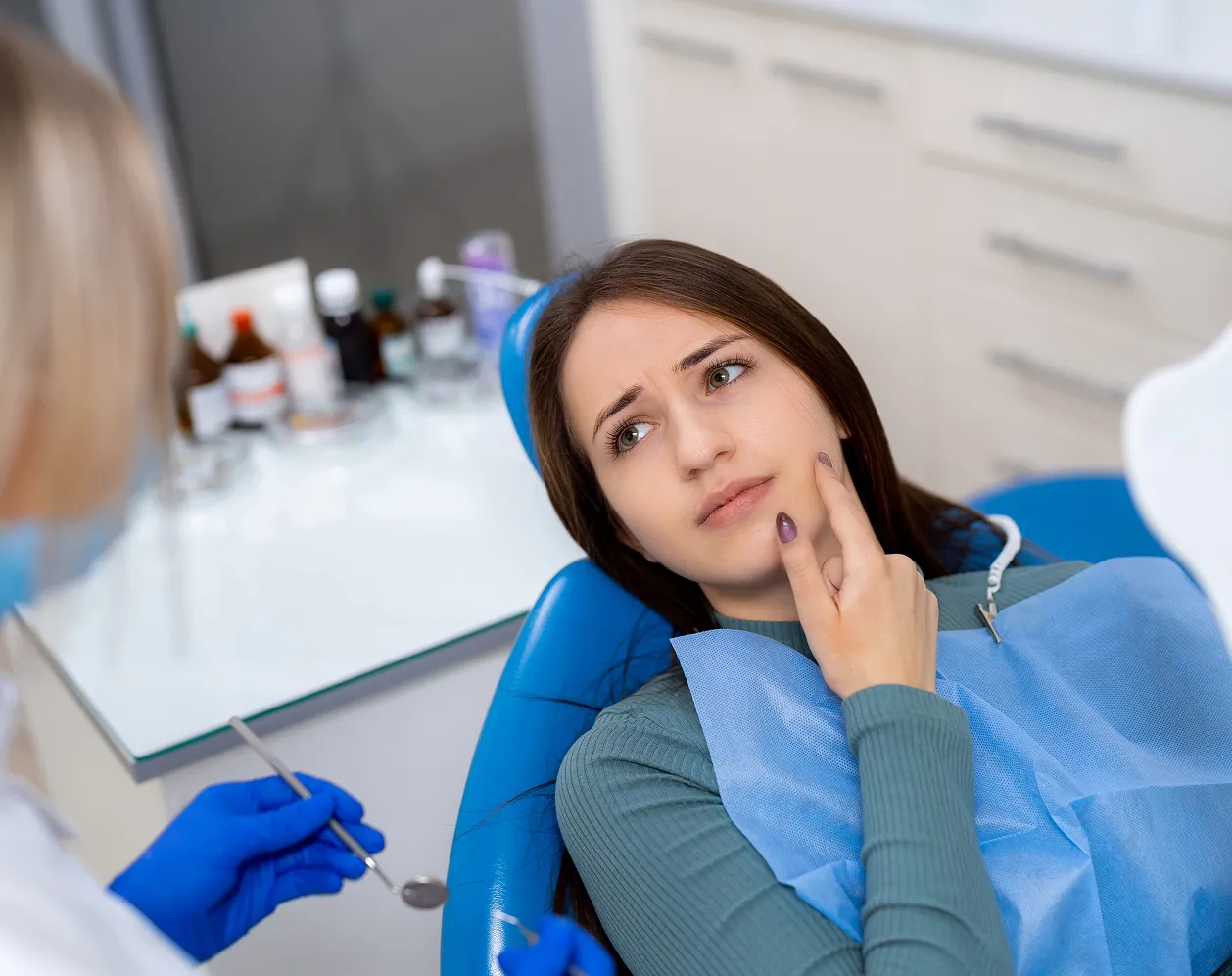 Young woman sitting in a dental chair looking worried and touching her cheek while a dentist wearing blue gloves holds dental tools.