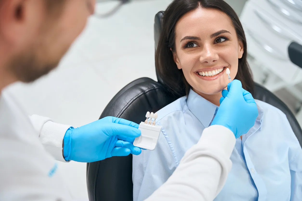 Dentist in blue gloves holding a tooth shade guide up to a smiling woman's teeth in a dental office.