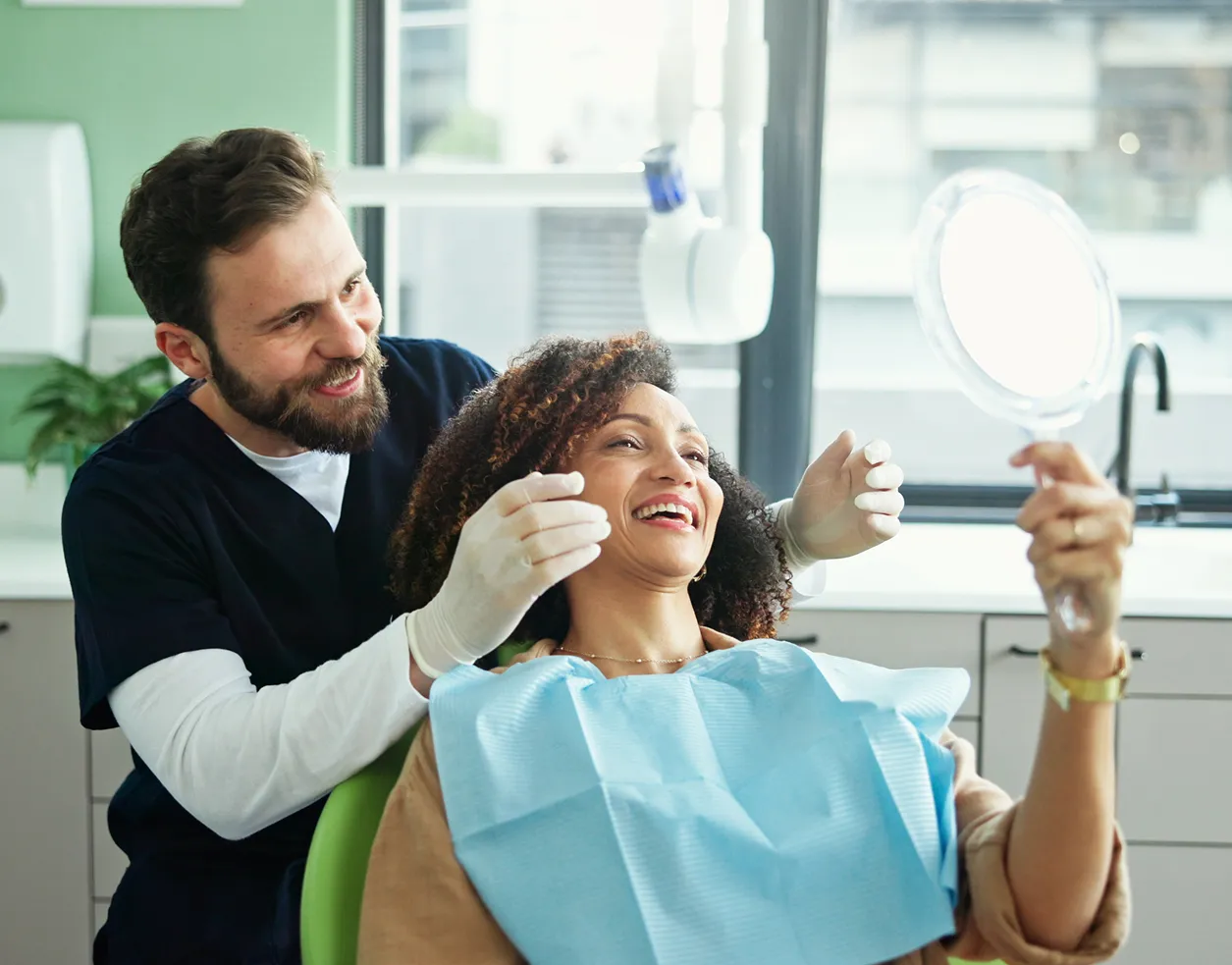 Smiling dentist in gloves examining happy female patient holding a mirror in dental clinic.