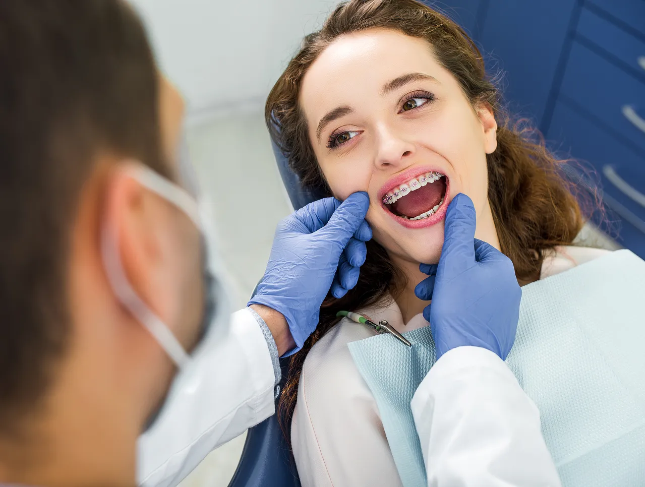 Dentist wearing blue gloves examining a young woman's braces in a dental clinic.