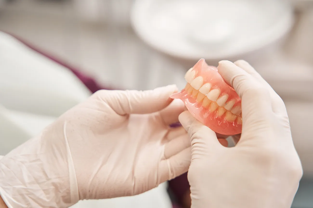 Hands wearing white gloves holding a set of upper dentures with pink gums and white teeth.