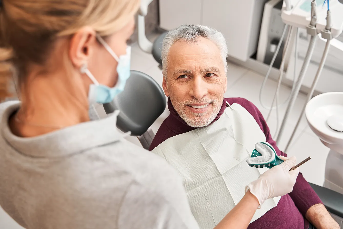 Dentist showing a dental mold to a smiling elderly man sitting in a dental chair.