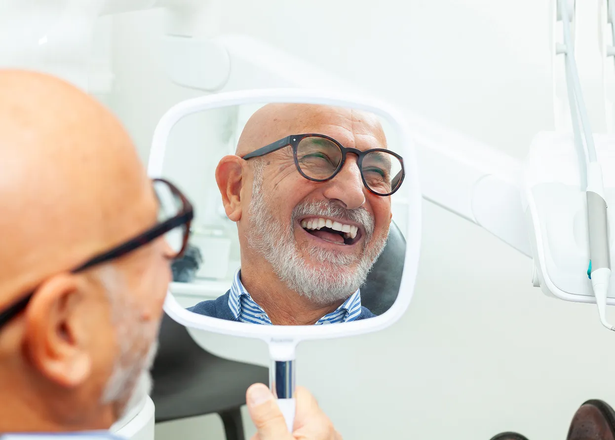 Smiling elderly man with glasses looking at his teeth in a handheld mirror at a dental clinic.