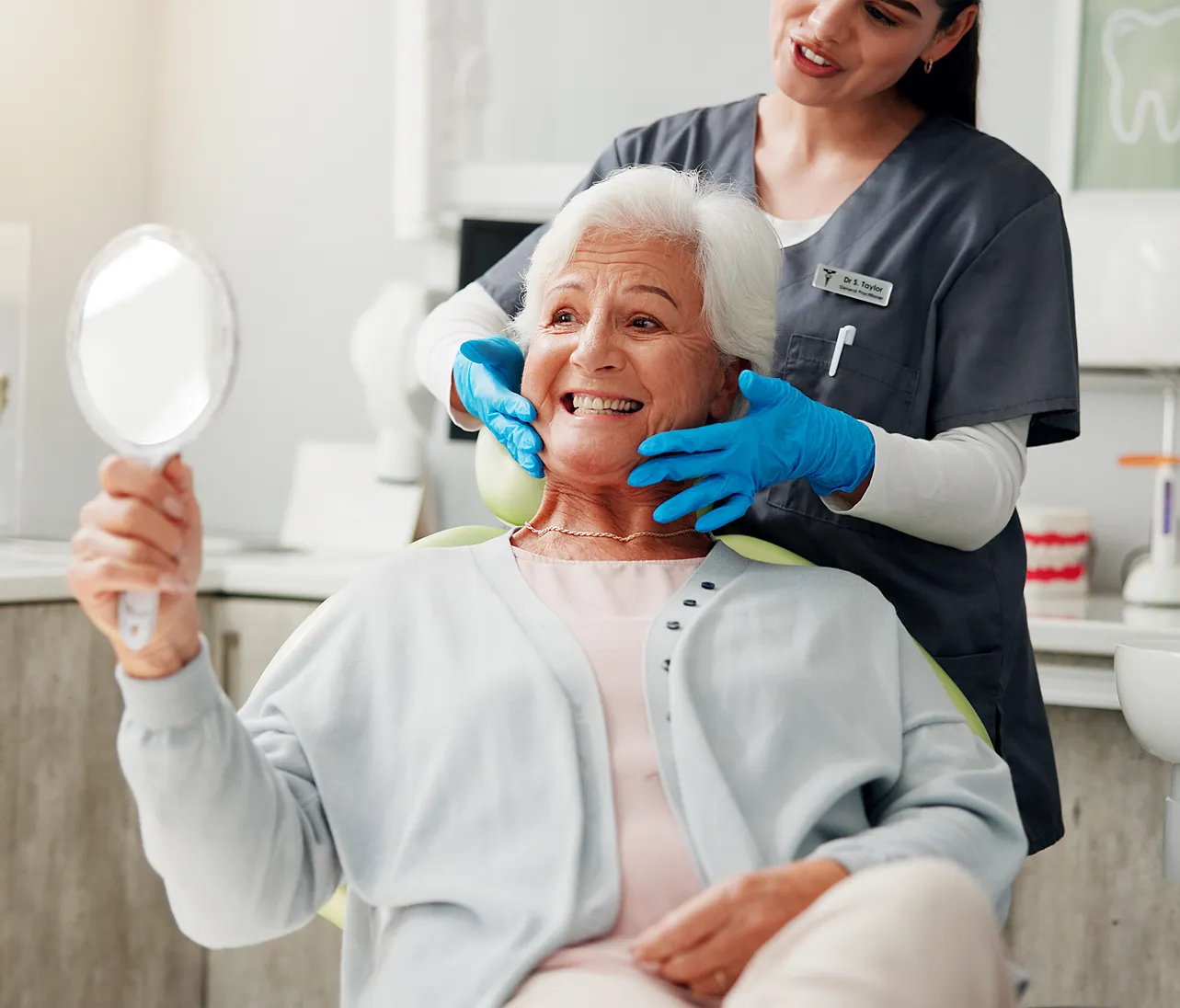 Dentist with blue gloves examining smiling elderly woman holding a hand mirror in a dental office.